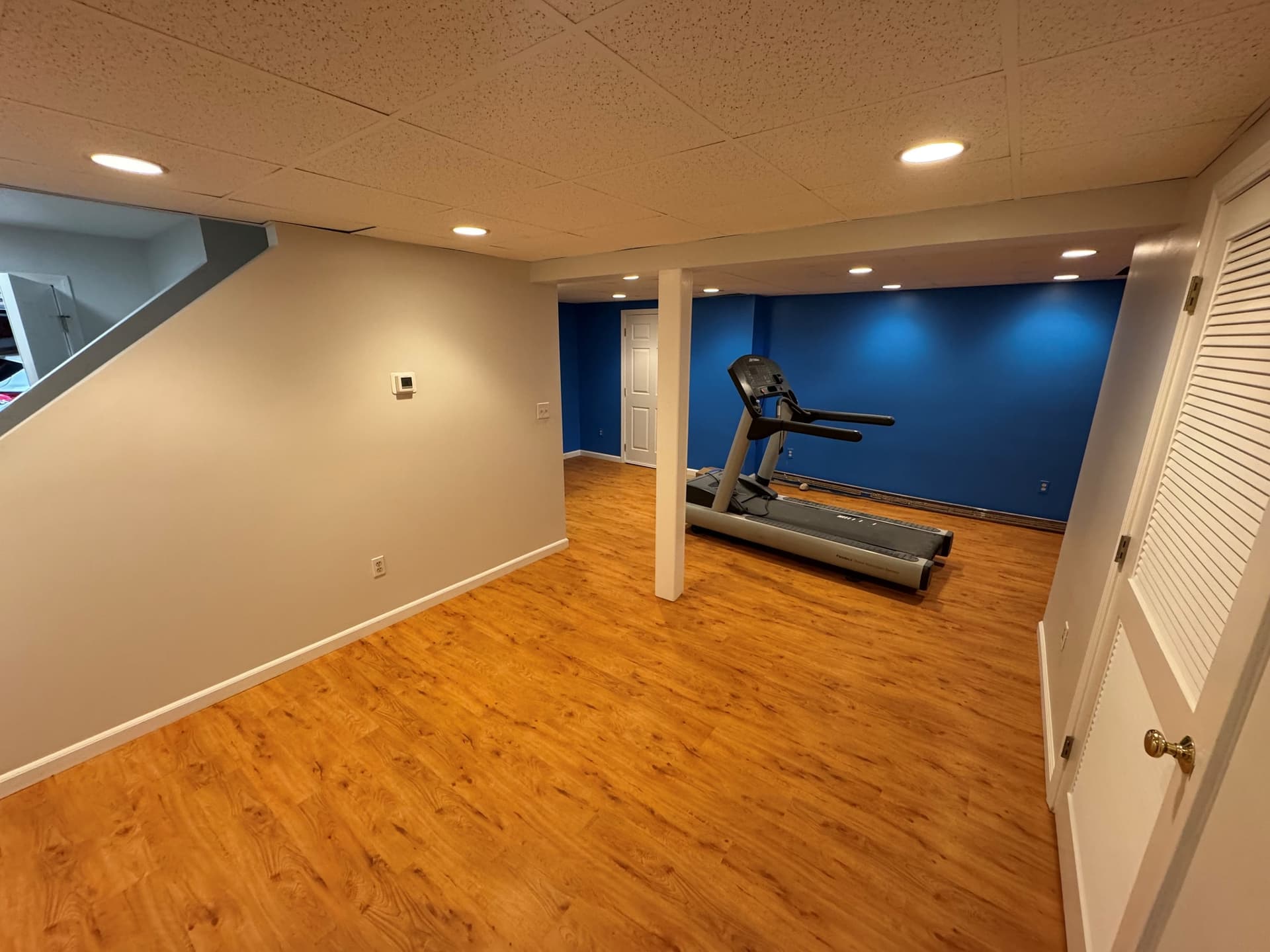 Completed glue-down Light Cherry vinyl flooring around a white support column and treadmill beside a blue basement wall